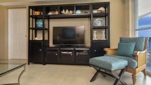 A living room with a dark entertainment unit, a flat-screen TV, a glass coffee table, and a blue upholstered armchair. - Luxurious Oceanview Condo in Cozumel (Cozumel, Centre)