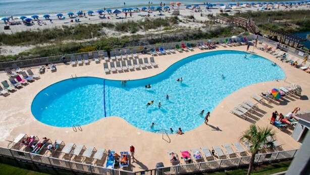 A pool with a curved edge, surrounded by lounge chairs and umbrellas, with a beach and ocean in the background.