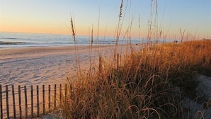 A beach with sand, grass, and a wooden fence.