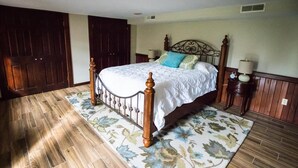 Une chambre avec un plancher en bois, un lit recouvert d’une courtepointe blanche, deux tables de chevet avec des lampes et une grande fenêtre.