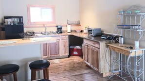 Eine kleine Küche mit Holzausstattung, einem integrierten Backofen, einer Kaffeemaschine und einem Toaster. Am Tresen stehen Hocker und an der Wand ein Metallregal.