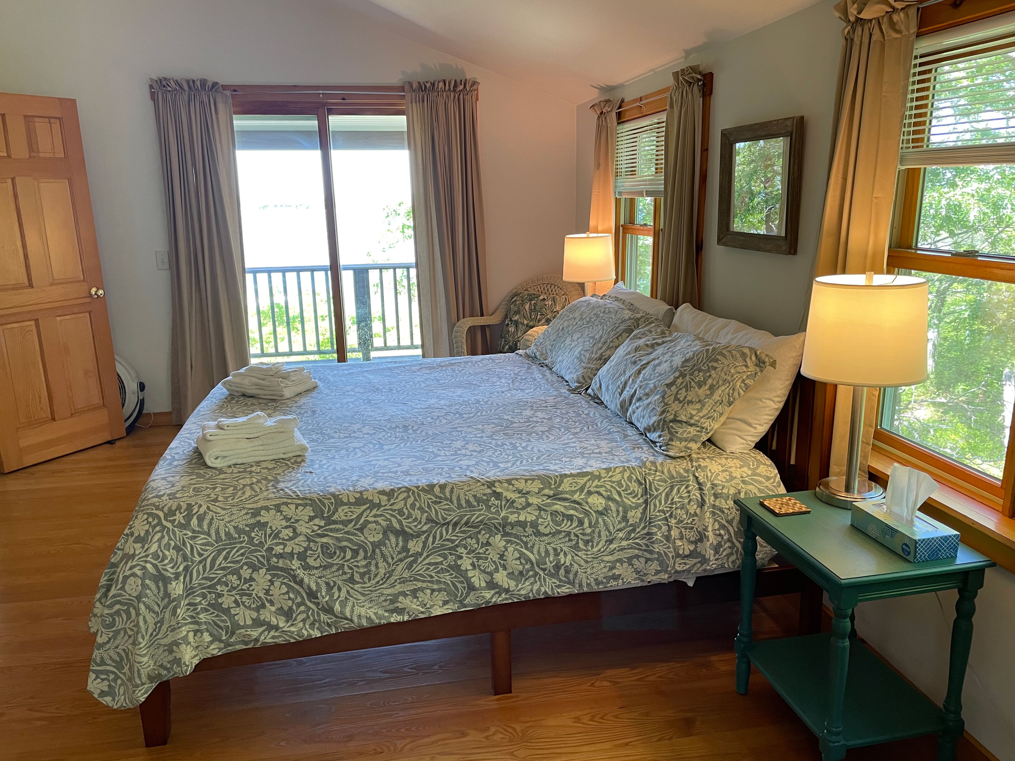 A bedroom with a bed, two bedside lamps, a bedside table, a wooden door, and a view of the outdoors.