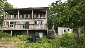 A two-story wooden house with a balcony, surrounded by dense greenery.