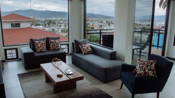 Ein modernes Wohnzimmer mit Sofa, Sessel und Couchtisch, mit großen Fenstern und Blick auf Stadt und Berge.