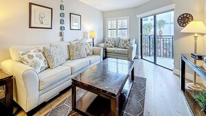 A living room with a beige sofa, patterned cushions, a glass coffee table, and a view of the outside balcony.