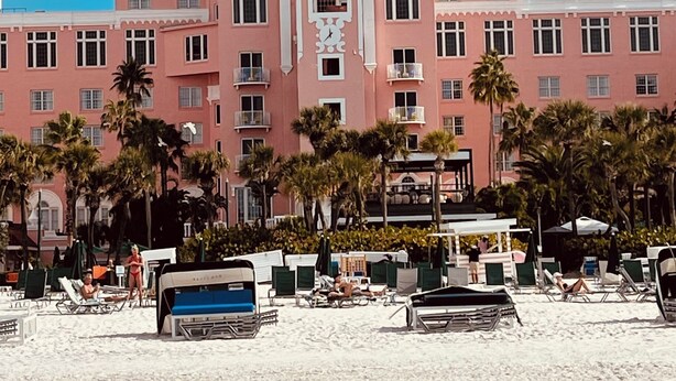 A large pink building with a central tower and two smaller towers, surrounded by palm trees and beach chairs.
