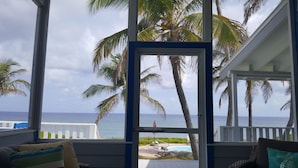 A view from a covered porch with a pool and palm trees, overlooking the ocean.