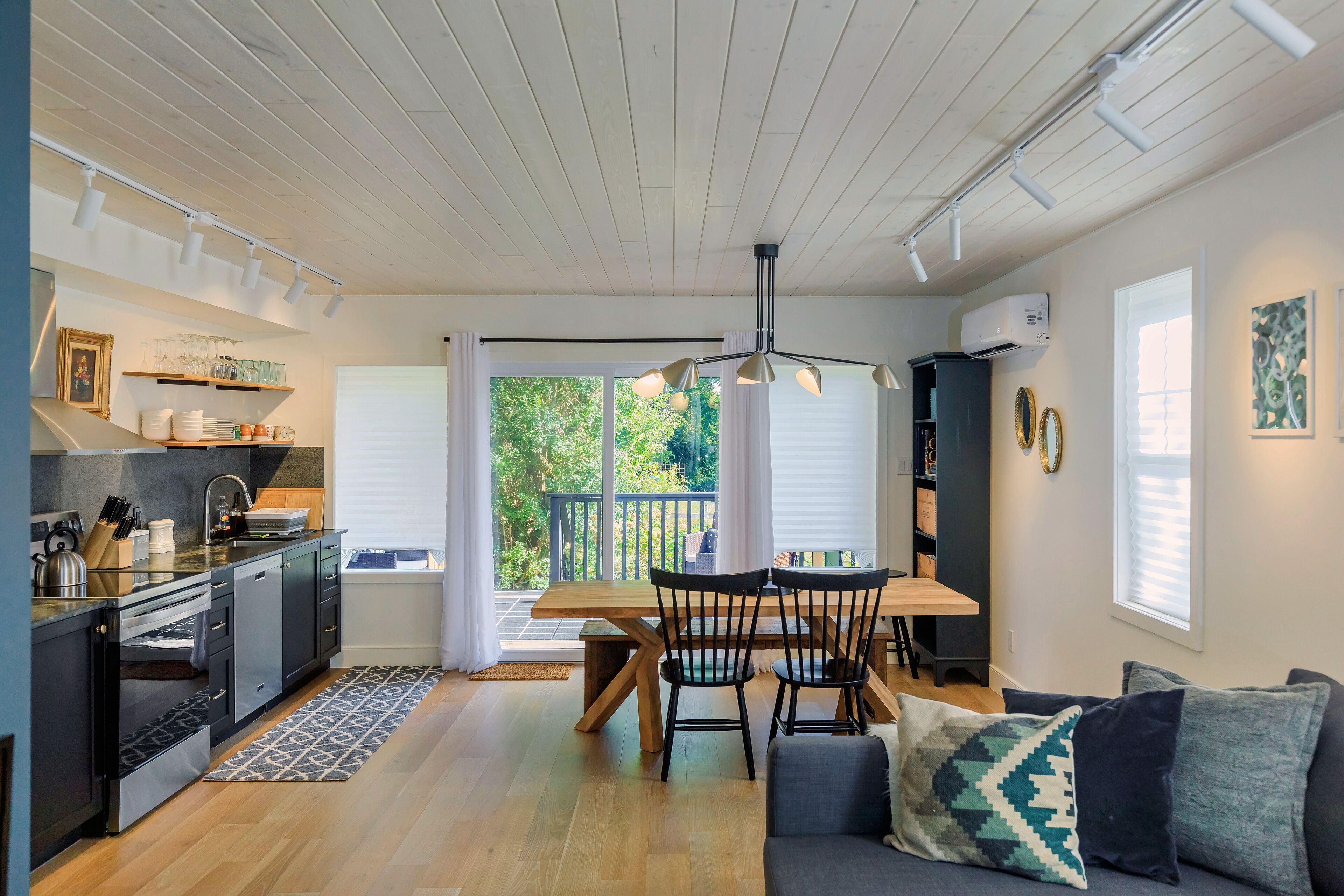 A modern kitchen with a dining area, wooden flooring, and a view of greenery outside.