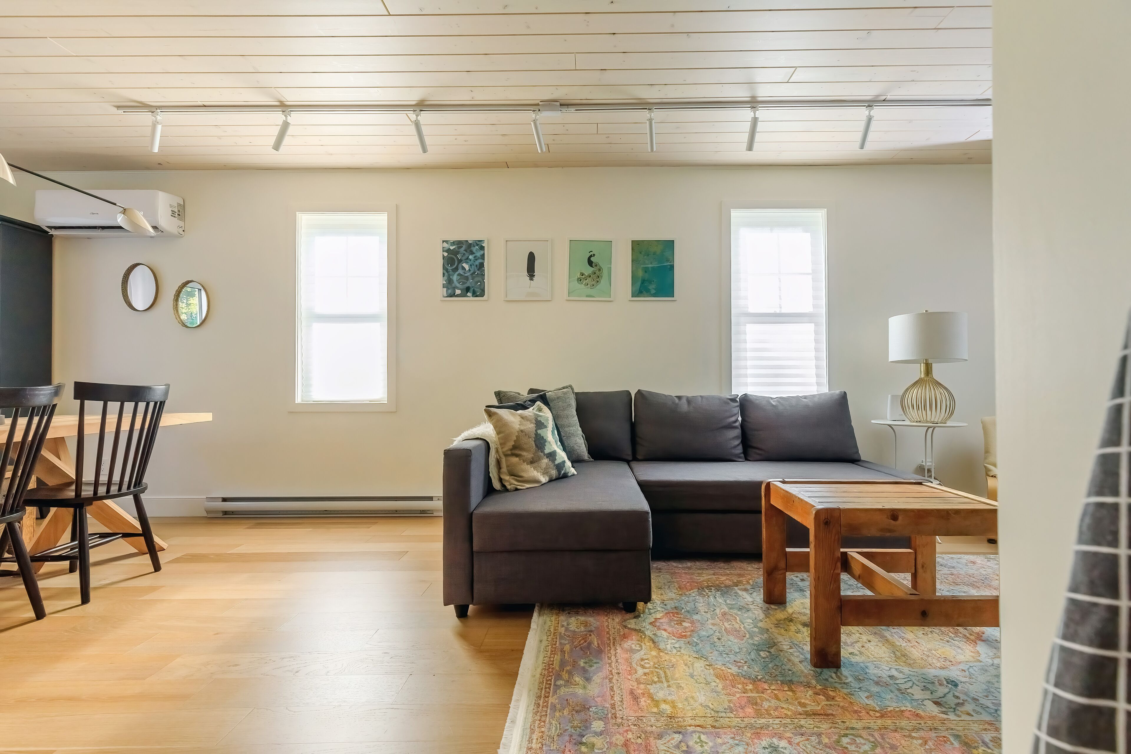A modern living room with a sectional sofa, a wooden coffee table, and a patterned rug. There is a dining area with a wooden table and chairs, and a small side table with a lamp. The room has large windows and a ceiling with exposed beams.