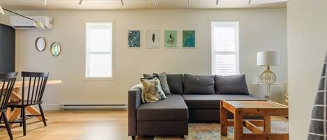 A modern living room with a sectional sofa, a wooden coffee table, and a patterned rug. There is a dining area with a wooden table and chairs, and a small side table with a lamp. The room has large windows and a ceiling with exposed beams.