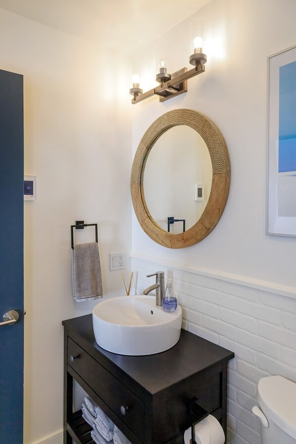 A bathroom with a round mirror, a white sink, and a dark vanity. - The Bellflower, Bloomfield (Prince Edward)