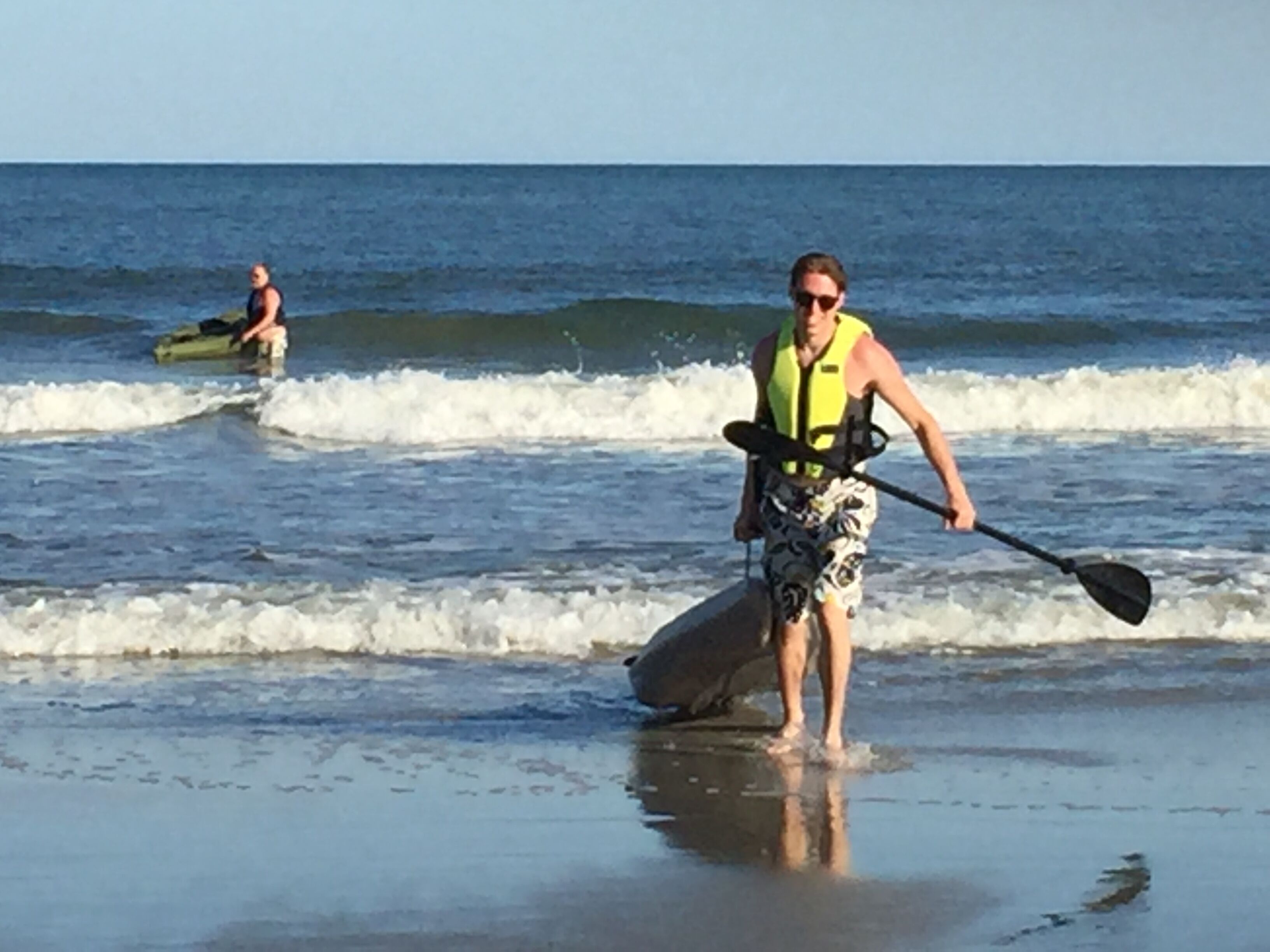 A person standing on a beach holding a paddleboard, with another person in the background on a kayak.