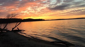 Ein Sonnenuntergang über einem ruhigen See mit sandigem Ufer und kahl stehenden Bäumen.