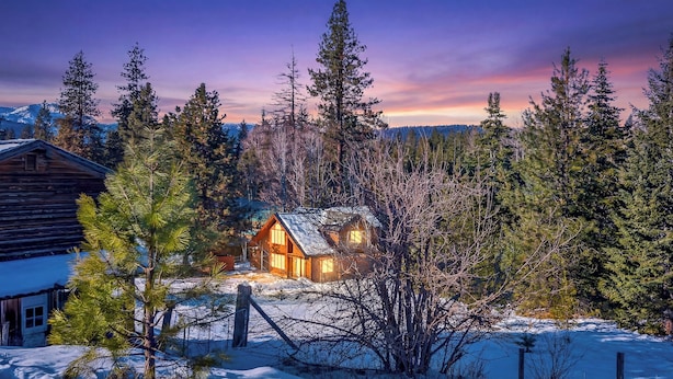 A log cabin with a snow-covered ground, surrounded by evergreen trees, and a mountain range in the background.