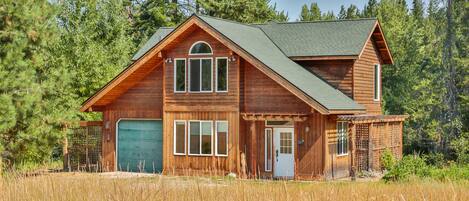 A two-story wooden cabin with a garage, surrounded by tall grass and trees.