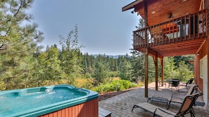 A hot tub with a wooden exterior, outdoor seating area with chairs and a table, and a view of a forested landscape.
