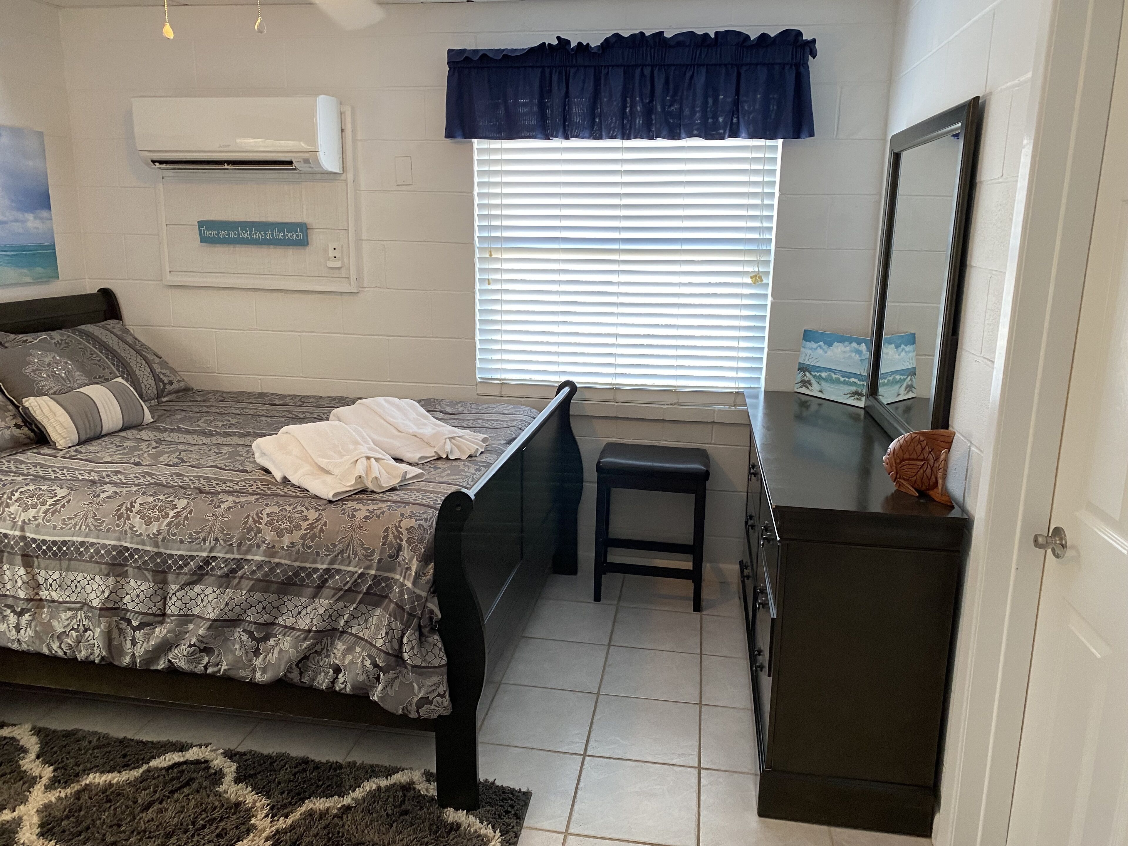 A bedroom with a bed, a small stool, a dresser, and a window with blinds.