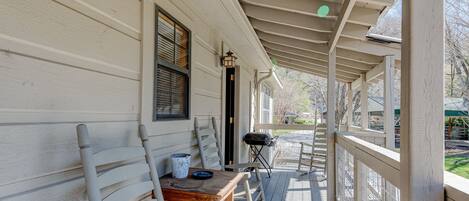A covered porch with a wooden table and chairs, a grill, and a window with blinds.