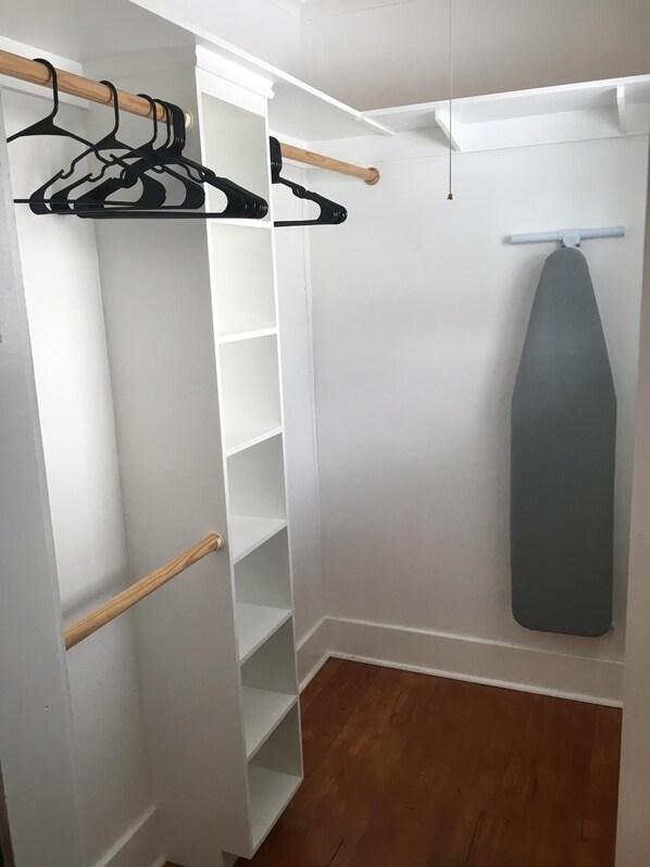 Un dressing accessible sans porte, doté d’étagères ouvertes, d’une tringle en bois et d’une planche à repasser suspendue.
