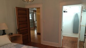 A bedroom with a wooden door, a bed with a blue and white quilt, a nightstand with a lamp, and a hallway leading to a bathroom with a mirror and a towel rack.
