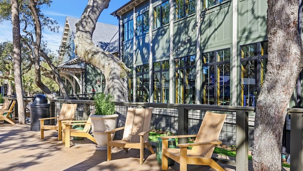 A wooden deck with chairs and a table, surrounded by trees and a building with large windows.