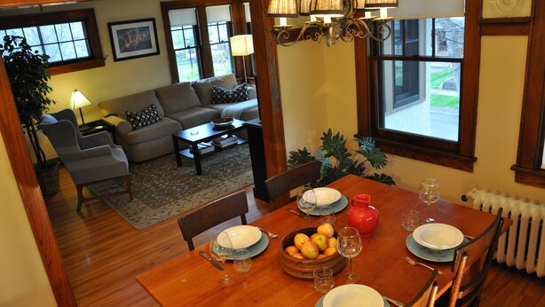 A dining area with a wooden table set for four, surrounded by chairs. There is a bowl of fruit on the table, and a red vase. The room has a view of a garden through large windows. - Fantastic duplex in the Historic District (Minneapolis)