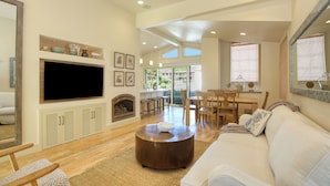 A spacious living room with a large flat-screen TV, a fireplace, a dining area with wooden furniture, and a large window offering a view of the outdoors. - Sunsetter in Avila Beach - 2 min walk to beach, located in Ocean 17 (Avila Beach)