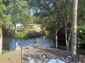 A lakeside patio with a fire pit, outdoor seating, and a view of a yellow building across the water.