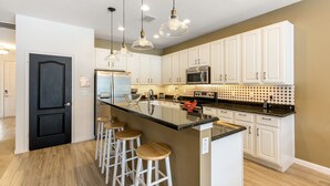 A modern kitchen with white cabinets, a central island with stools, pendant lighting, and a stainless steel refrigerator.