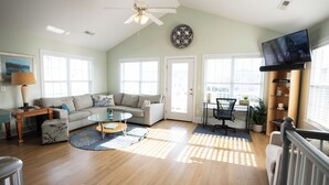 A spacious living room with a large sofa, a glass coffee table, and a TV mounted on the wall. - Your Perfect Beach House (Surf City)