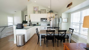 A modern kitchen with white cabinets, a central island, and a dining area with black chairs. - Your Perfect Beach House (Surf City)