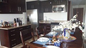 A kitchen with dark wooden cabinets, a stainless steel refrigerator, a microwave, and a dining table set with chairs and a vase of white flowers.