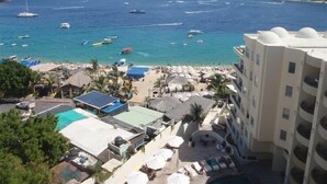 A coastal hotel with a pool, beach umbrellas, and a view of the sea with boats.