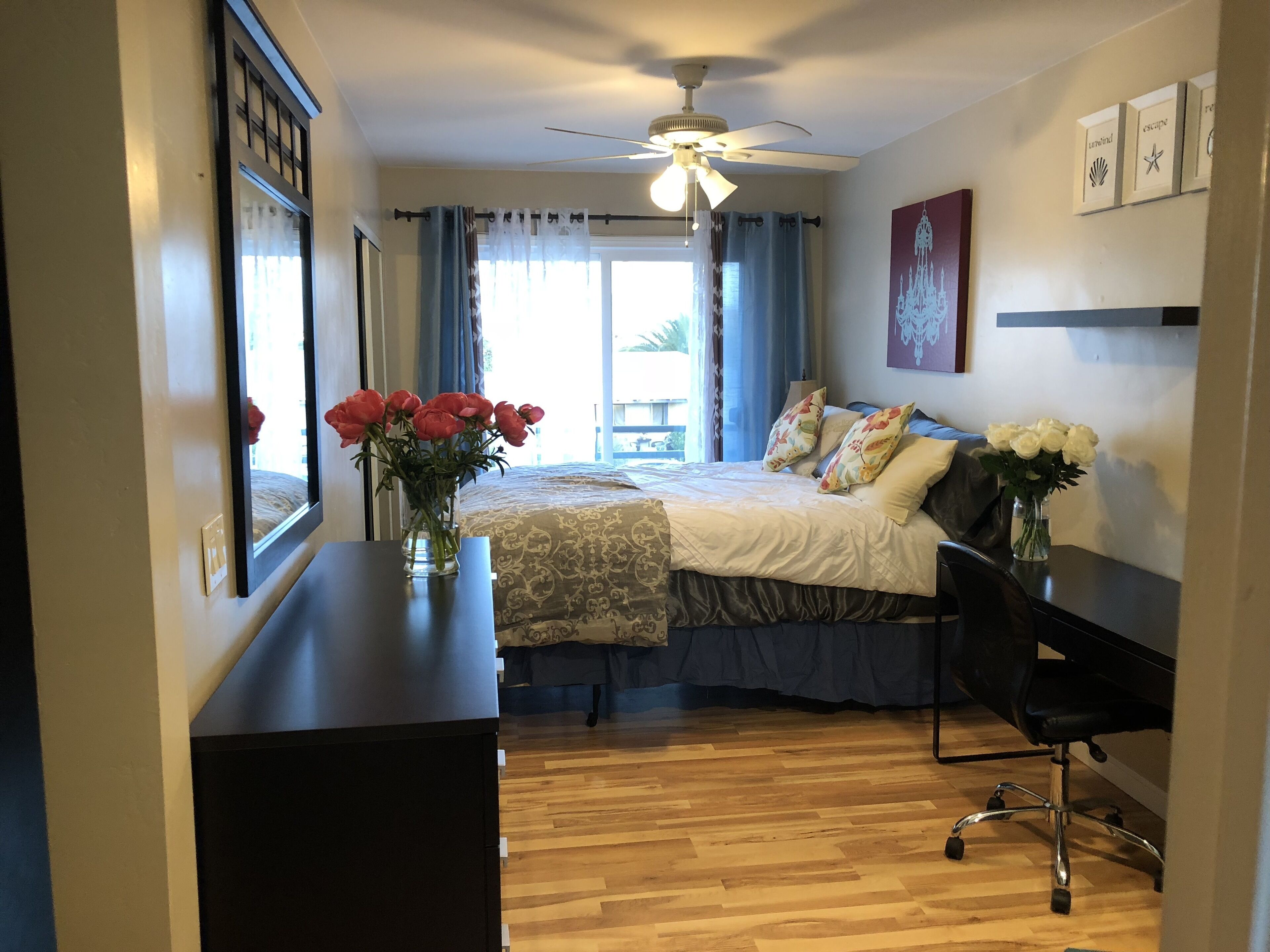 A bedroom with a bed, a desk, a chair, a ceiling fan, and a window with curtains.