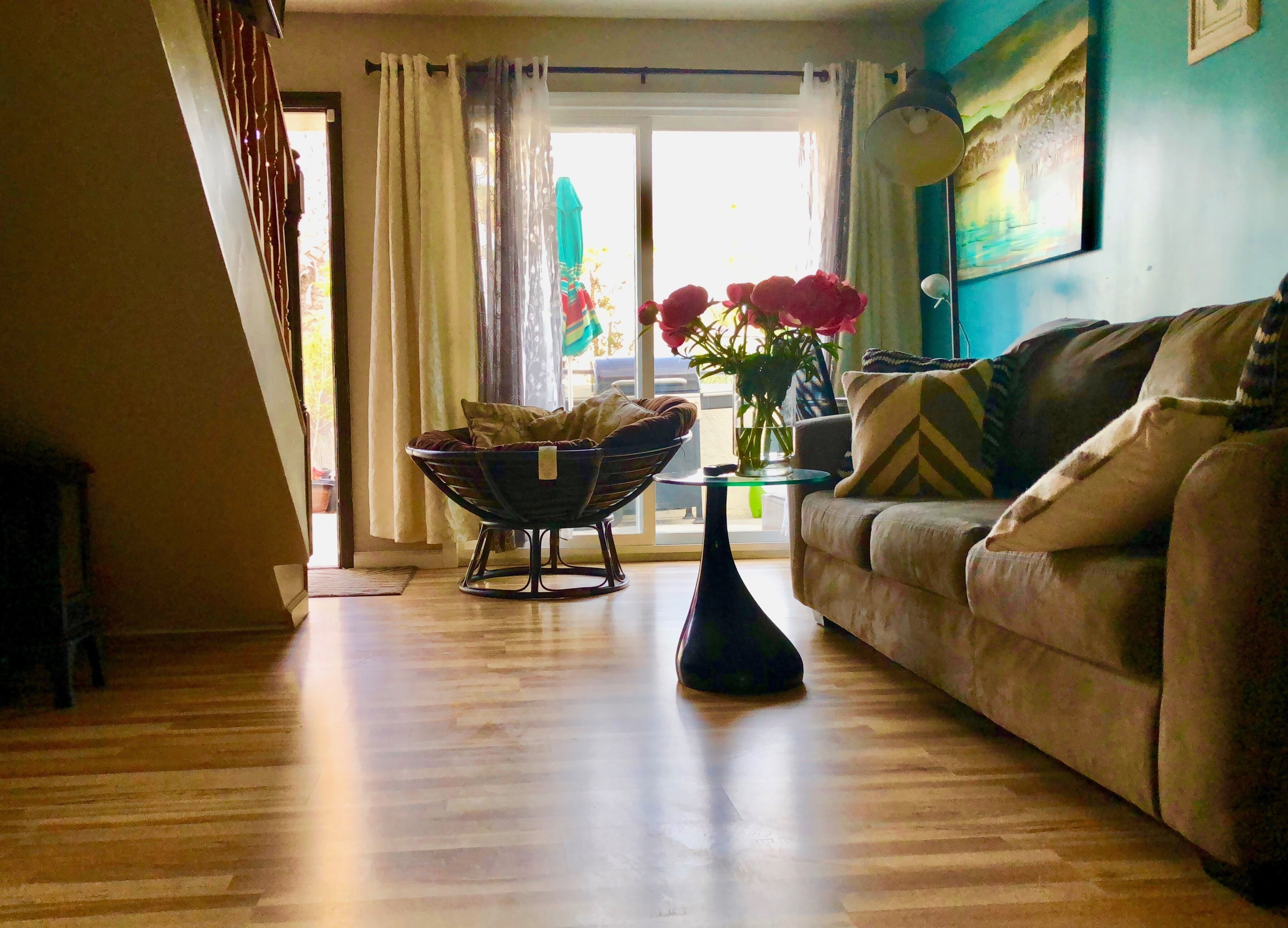 Una sala de estar con un sofá, una mesa redonda para café y una lámpara de pie. Hay una ventana con cortinas y un cuadro en la pared.