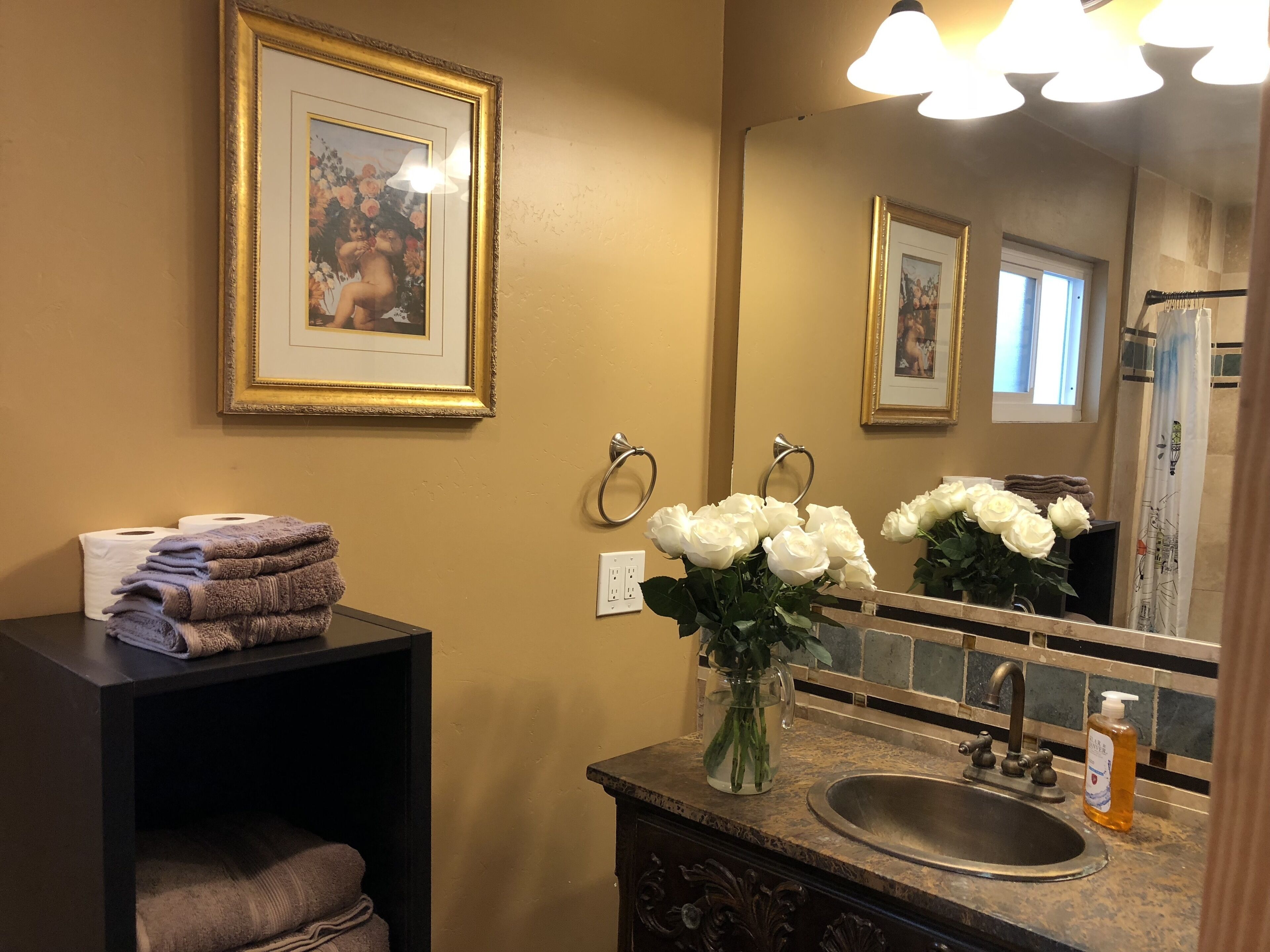 A bathroom with a sink, mirror, framed picture, towels, and a vase with flowers.
