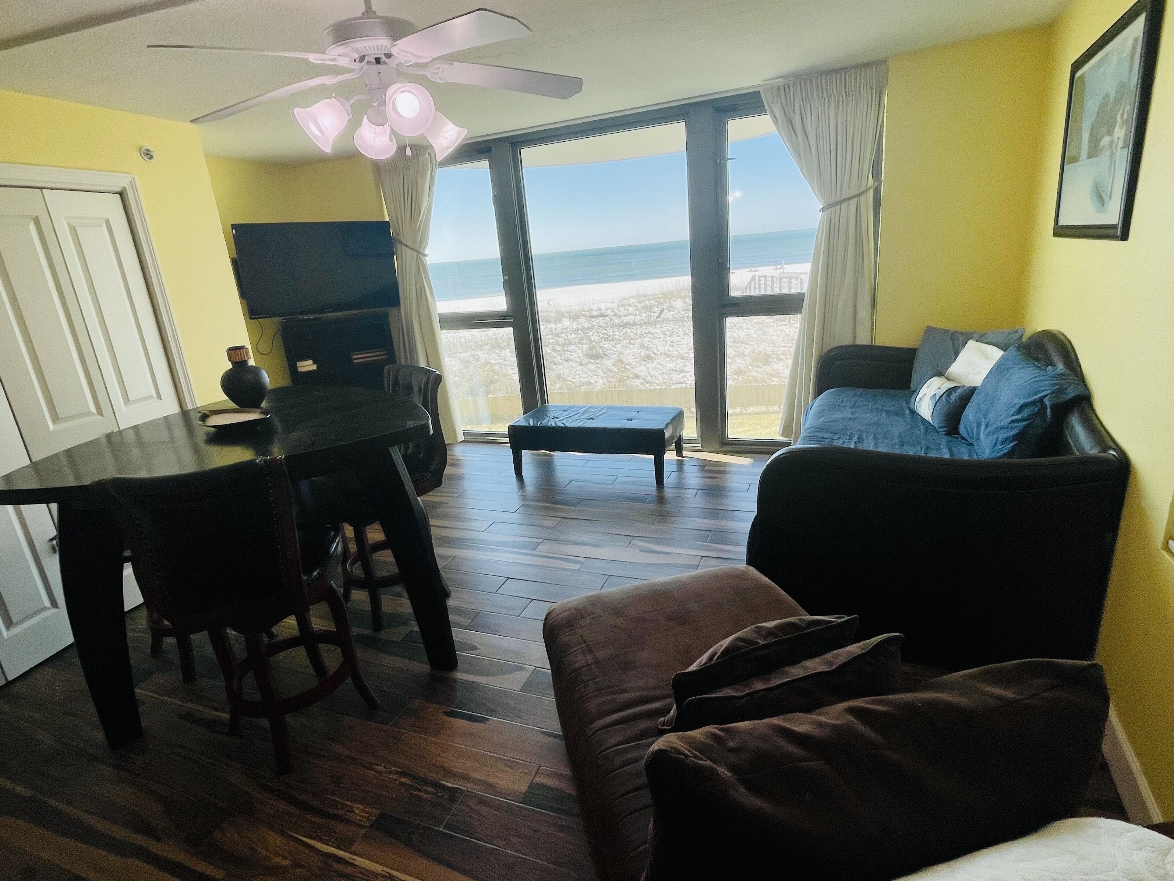 A living room with a sofa, a coffee table, and a TV. There is a balcony with a view of the beach.
