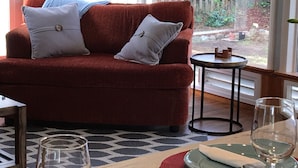 A cozy living room with a red sofa, two grey pillows, a small round table, and a glass of wine. - NEW! Tranquil Oceanfront Gem with Hot-tub 3 BDRM + Den, 3 BATH (Waldport)