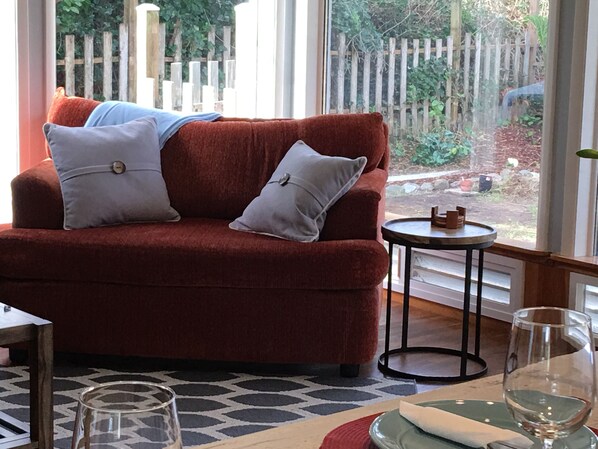 Una sala de estar acogedora con un sofá rojo, dos almohadas grises, una mesita redonda y una copa de vino.