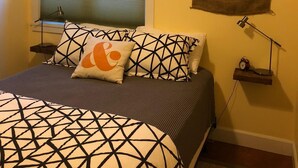 Een slaapkamer met een bed, kussens, een raam, een wanddecoratie en een nachtkastje met een lamp.
