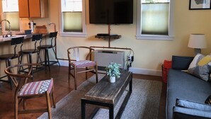 Een woonkamer met een bank, een salontafel en een televisie. Achtergrond toont stoelen en een keuken.