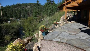 Un sendero de piedra que lleva a una construcción de madera con base de piedra, rodeada por un paisaje natural con un río y un bosque denso.