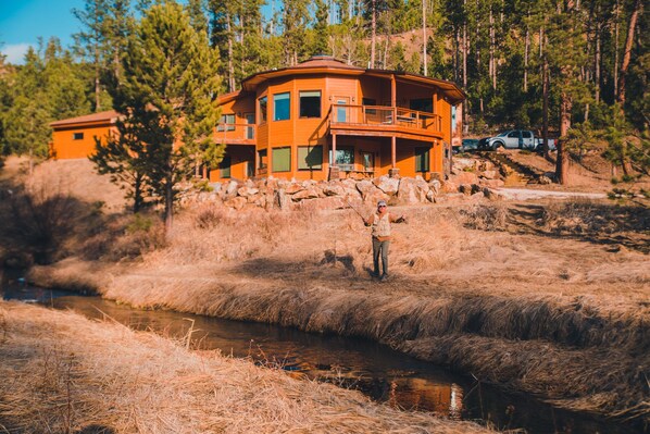 A large, multi-story wooden cabin with a balcony, surrounded by a rocky landscape and a stream. - Spring Creek Splendor. The Perfect Location. Private Lower Level Apartment. Fun! (Hill City)