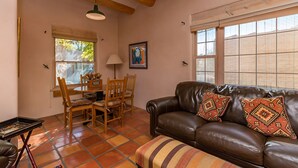 A living room with a brown leather sofa, patterned cushions, a dining table with chairs, and a window with blinds. - Favorite Downtown Adorable Adobe ~ 10 min walk to Plaza (Santa Fe)