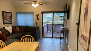 Una sala de estar con un sofá, una mesa de comedor de madera con sillas, un ventilador de techo y un ventanal corredero que da a una terraza exterior.
