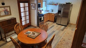 A dining area with a wooden table and chairs, a kitchen with wooden cabinets and appliances, and a patterned rug on the floor. - Escape to Cool Twin Pines! (Pine)