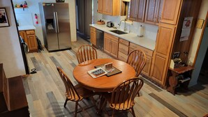A modern kitchen with wooden cabinets, a stainless steel refrigerator, and a dining area with a wooden table and chairs. - Escape to Cool Twin Pines! (Pine)