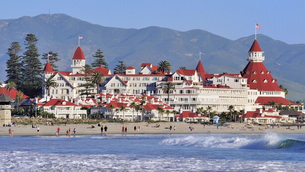 A coastal hotel with multiple red-roofed buildings, a beach in the foreground, and mountains in the background.
