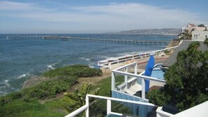 A coastal view with a long pier extending into the sea, a building with a balcony overlooking the ocean, and lush greenery in the foreground.