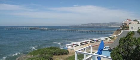 A coastal view with a long pier extending into the sea, a building with a balcony overlooking the ocean, and lush greenery in the foreground.